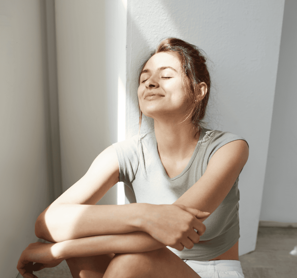 Una mujer está sentada en el suelo junto a una pared blanca, con los ojos cerrados y una leve sonrisa, y la luz del sol le ilumina la cara y los brazos.