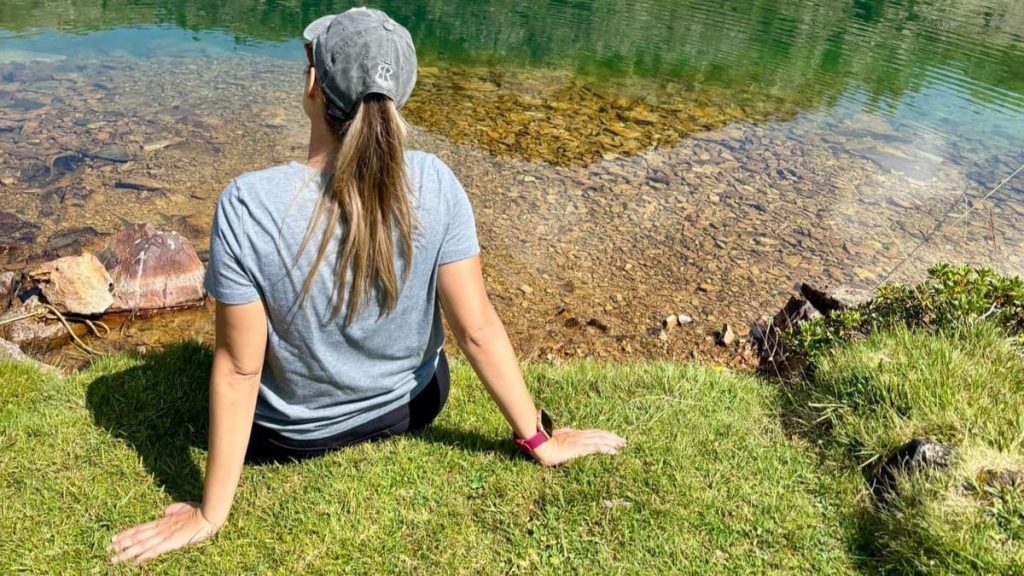 Una persona con camisa y gorra grises está sentada en el suelo cubierto de hierba junto a la orilla de un lago claro y rocoso, mirando hacia el agua.