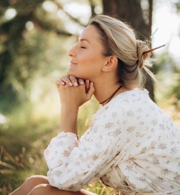 Una mujer de pelo rubio est&aacute; sentada al aire libre, con los ojos cerrados, las manos juntas bajo la barbilla y un ligero vestido de flores. La luz del sol se filtra entre los &aacute;rboles del fondo.