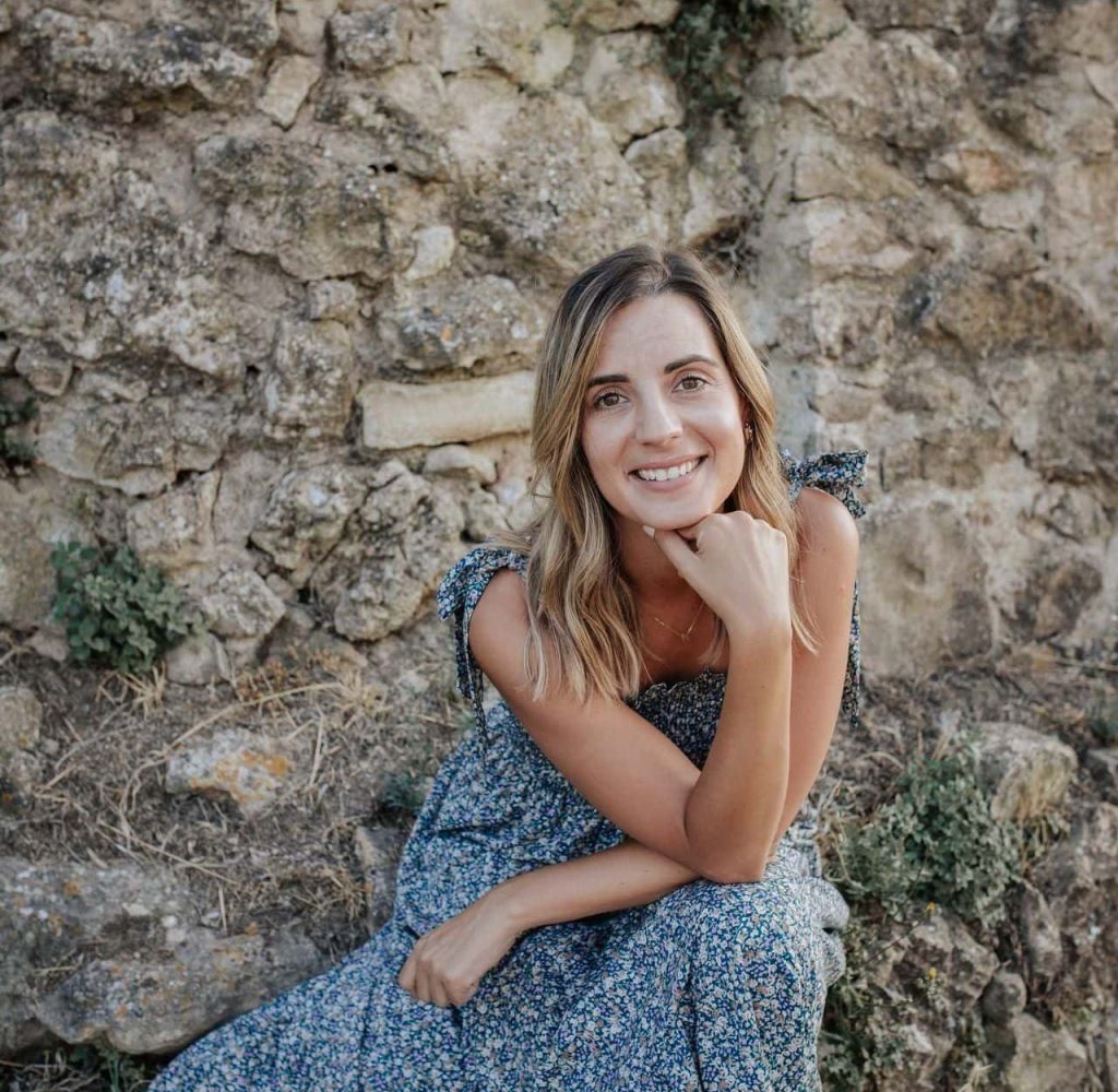 Mujer con vestido azul de flores sentada frente a un muro de piedra, sonriendo a la cámara con la mano apoyada bajo la barbilla.